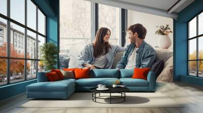 Young happy couple having fun talking laughing relaxing at home on couch, boyfriend embracing girlfriend telling funny joke sitting on sofa, humor in relationships, enjoying weekend together. Wall mural