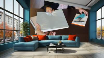 Woman is handing over a white file in direction camera while sitting at the desk in the office. Wall mural