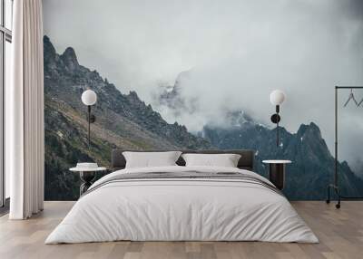 Dark atmospheric surreal landscape with dark rocky mountain top in low clouds in gray cloudy sky. Gray low cloud on high pinnacle. High black rock with snow in low clouds. Surrealist gloomy mountains. Wall mural