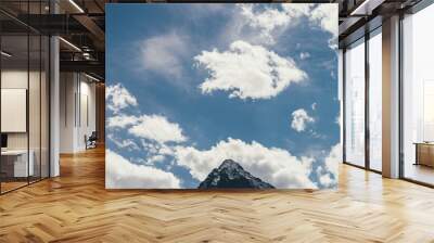 Minimalist cloudscape with pointy peak in cloudy blue sky. Minimal sky landscape with cirrus clouds above peaked top with snow. Beautiful mountain scenery with sharp rock under spindrift clouds. Wall mural