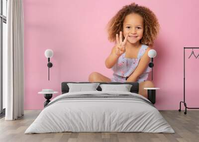 Beautiful little girl sitting on the floor counting three with fingers over pink background. Wall mural