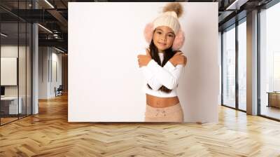 Cute smiling girl wearing stylish earmuffs hugging herself on white background Wall mural