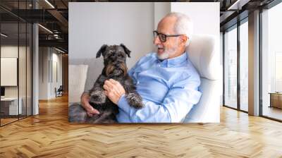 Happy Grandpa is sitting at chair in home with dog Jack Russell Terrier on his legs. Senior man 70-75 years old smiling at camera. Light window on background. Сoncept of happy old age. Wall mural