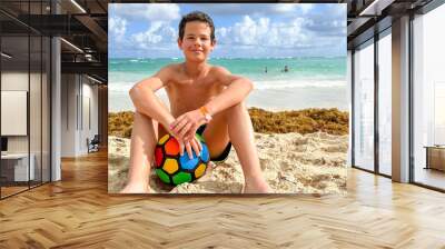 Portrait of a cute European teen boys wearing swimming shorts. The boy sitting on the sand holding a ball. Wall mural