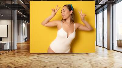 Young happy brunette woman wearing swimsuit over isolated yellow background doing peace symbol with fingers, smiling cheerful showing victory. Wall mural