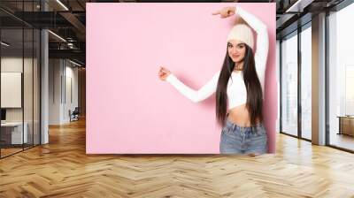 Young woman wearing winter hat over isolated pink background pointing fingers to the side Wall mural