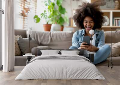 Excited happy young black woman holding smart phone device sitting on sofa at home - Happy satisfied female looking at mobile smartphone screen  - Technology concept Wall mural