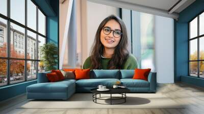 A close up head shot portrait of a preppy, young, beautiful, confident and attractive Indian Asian woman in a green sweater and spectacles in a classroom or office. She is smiling happily. Wall mural