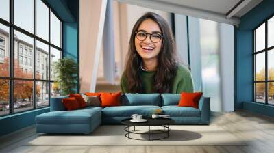 A close up head shot portrait of a preppy, young, beautiful, confident and attractive Indian Asian woman in a green sweater and spectacles in a classroom or office. She is smiling happily. Wall mural