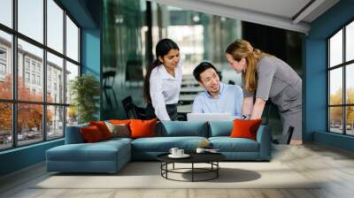 A diverse multi-racial team of three colleagues businesspeople have a relaxed discussion around a laptop notebook computer in an office or coworking space. They are all smiling and talking animatedly. Wall mural