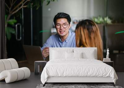 A pair of diverse team mates have a business meeting and discussion together to collaborate. A Korean man is sitting at a table in a trendy coworking space office and talking to his female companion. Wall mural