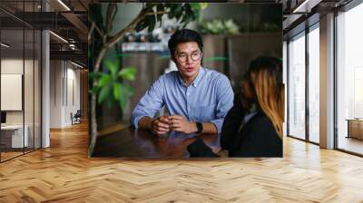 A pair of diverse team mates have a business meeting and discussion together to collaborate. A Korean man is sitting at a table in a trendy coworking space office and talking to his female companion. Wall mural