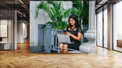 An attractive and confident Indian Asian woman seated in a bench during the day while workong on her laptop. She is taking a short break and checking the messages on her phone. Wall mural