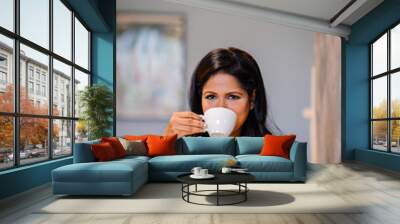 An image of a confident and successful Indian Asian Desi woman sitting at a table and wearing a professional black dress for her attire.She is drinking her coffee while waiting for her workmates. Wall mural