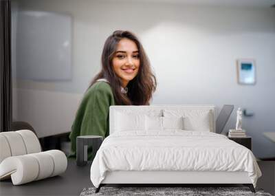 Portrait of a beautiful, young and intelligent-looking Indian Asian woman student wearing a white shirt and green tracker smiling as she works on her laptop in a university classroom. Wall mural