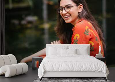 Portrait of a young, attractive Indian Asian woman in a traditional Indian orange dress smiling as she sits on a step in the city during the day. Wall mural