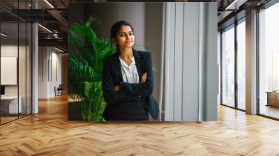 Portrait of a young Asian Indian businesswoman in a meeting room standing by the window, smiling with her arms crossed. She looks optimistic, happy and confident. Wall mural