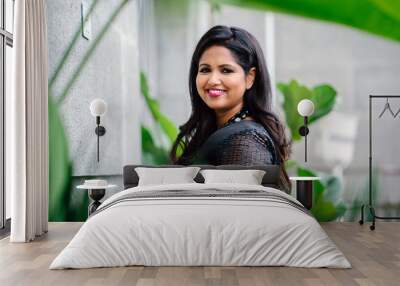 Portrait of a youthful but confident and mature Indian woman. She is dressed in an elegant and classy black dress. She is standing in a hallway next to some green plants while striking a radiant smile Wall mural