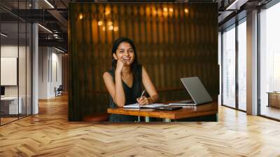 Portrait of Indian lady writing in a notebook in a cosy cafe with a laptop nearby Wall mural