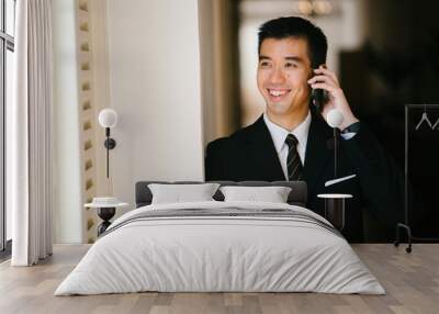 Portrait of young handsome man standing in a well lit corridor. He is wearing formal business attire and talking and smiling on his phone.  Wall mural