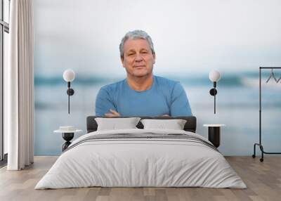 Middle shot of a senior man in blue t-shirt with arms crossed on the chest. Person looking to the camera with a light smile. View against blurry background of sea and sky Wall mural