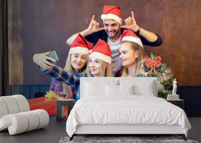 Happy young friends wearing Santa claus hat enjoying Christmas party taking selfie with funny faces at home sharing it on social media Wall mural