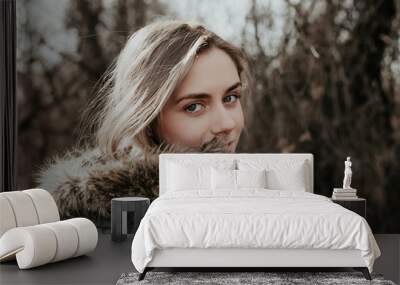 Beautiful young woman with blonde hair, wide eyebrows and blue eyes looking at camera over her shoulder. Girl posing at camera in green jacket. Wall mural