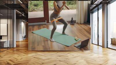 Close up of woman in activewear doing squats with fitness elastic band on mat at home Wall mural