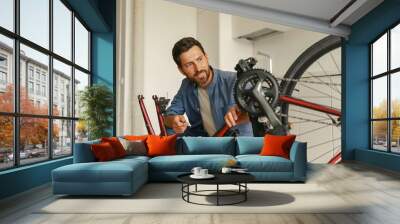 Handsome smiling man in casual clothing repairing bicycle itself at home Wall mural