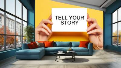 Female hands hold card paper with text Tell Your Story on a yellow background. Business and finance concept Wall mural