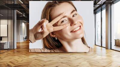 Hey smile you. Young beautiful girl smiles and shows peace victory gesture as fooling around on a white isolated background. A woman points to an idea, a place for advertising. Positive cute brunette Wall mural