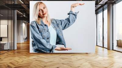 Smiling blonde girl shows big size with her hands on a gray background in the studio. A woman in casual clothes is happy with the measure. Place for advertising. Wall mural