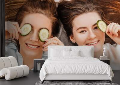 Two girls make homemade face and hair beauty masks. Cucumbers for the freshness of the skin around the eyes. Women take care of youthful skin. Girlfriends laugh at home lying on the floor on pillows. Wall mural