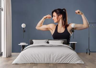 Portrait of a happy elegant caucasian brunette woman showing her biceps muscles on the arms in black clothes and fitness over gray background, smiling and looking at camera Wall mural