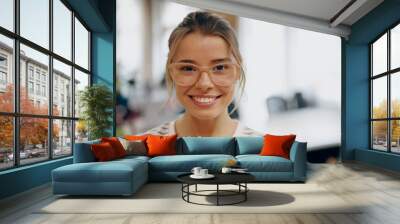 Portrait of young female manager in eyeglasses standing on office background and looks camera Wall mural