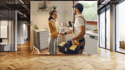 Professional repairman in uniform shaking hands with woman while standing at home kitchen Wall mural