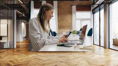 Smiling young business woman working on laptop and use phone while sitting in stylish office  Wall mural