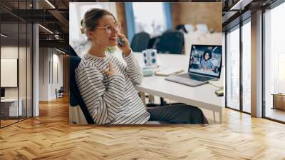 Young smiling female entrepreneur talking by phone with client while sitting in modern office Wall mural