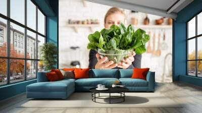 Young blond smiling woman holding a bowl of fresh spinach in the kitchen Wall mural