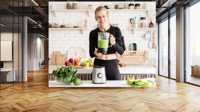 Young blond smiling woman making green smoothie at home kitchen Wall mural