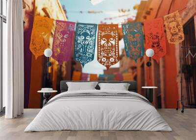 Colorful papel picado banners featuring skull designs hang between buildings, creating a festive atmosphere in a lively street during Dia de los Muertos Wall mural