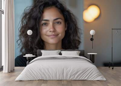 A woman with curly hair and a black shirt smiles warmly indoors, lit by soft, natural light Wall mural