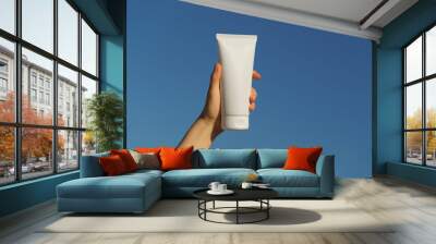 Female hand holding a white mockup tube of cream on a blue sky background. Concept of natural moisturizing, nourishing and sun protection cosmetics for skin, beauty and spa. Wall mural