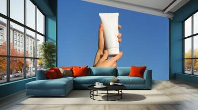 Female hands holding a white mockup tube of cream on a blue sky background. Concept of natural cosmetics for body or face, daily care, protection with spf. Wall mural