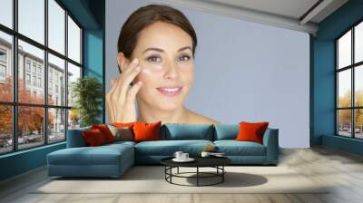 Beautiful young woman applying face cream to her cheek bone in a beauty  skincare or cosmetics concept  on grey with copy space Wall mural