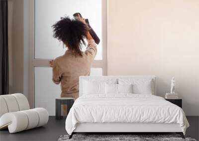 A curly-haired mixed-race girl in casual clothes wipes the window with a rag. A young African woman cleans a house or apartment. Wall mural