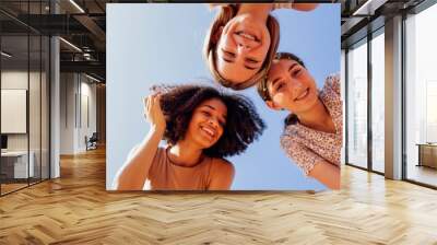 Funny multiracial smiling teenager gilrs standing and looking down at camera. Group of happy cheerful female friends in casual clothes having fun together outdoors. Low angle view. Copy space. Wall mural