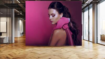 Beautiful fashionable girl haired with long curly hair posing with a pink bow. The girl in the studio on a pink background. Fashion, beauty. Bright makeup. Cosmetics for hair, healthy hair. Wall mural