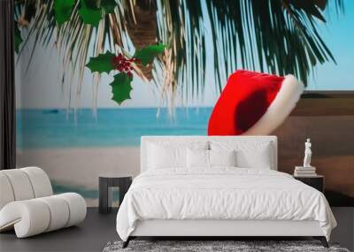 A red Santa hat is placed on a beach lounger, and palm leaves hanging from above in front of blue ocean water Wall mural