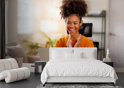 Female digital nomad smiling happily as she types on her laptop on sofa. Afro haired woman dressed in casual clothing and working online in a relaxed environment. Wall mural